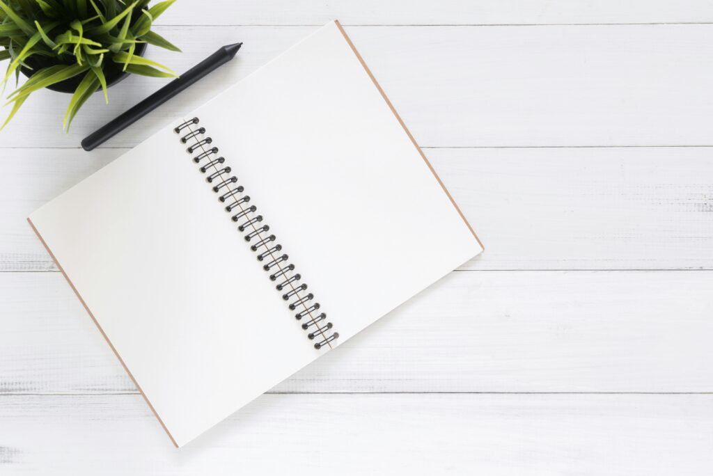 Open spiral notebook on white desk with black pen and green plant. Ideal for planning.